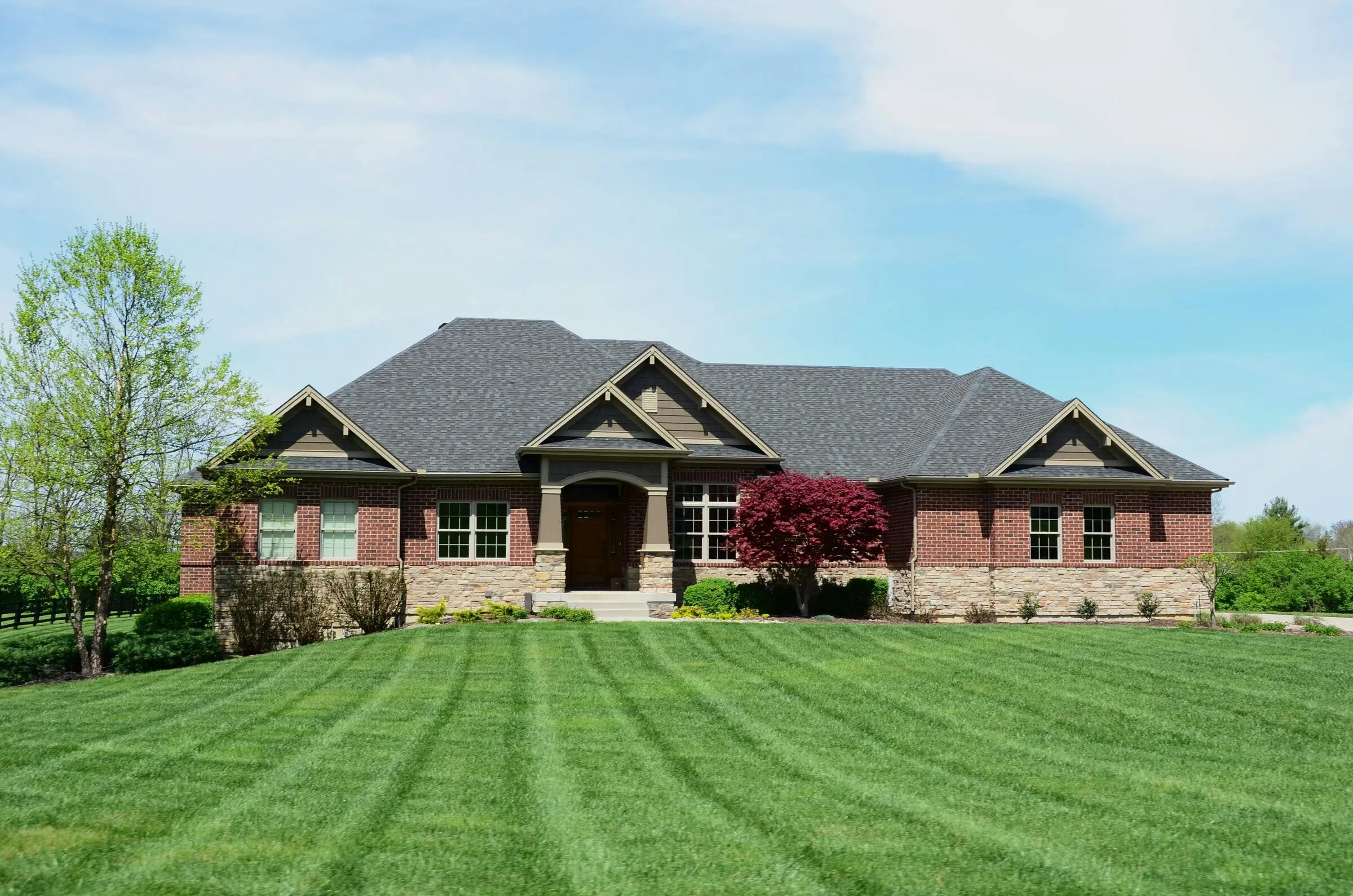 A single-storey brick and stone ranch house behind a wide, freshly mowed lawn with light and dark mowing stripes, a red Japanese maple by the porch and a pale blue sky with thin cloud filling the upper half.