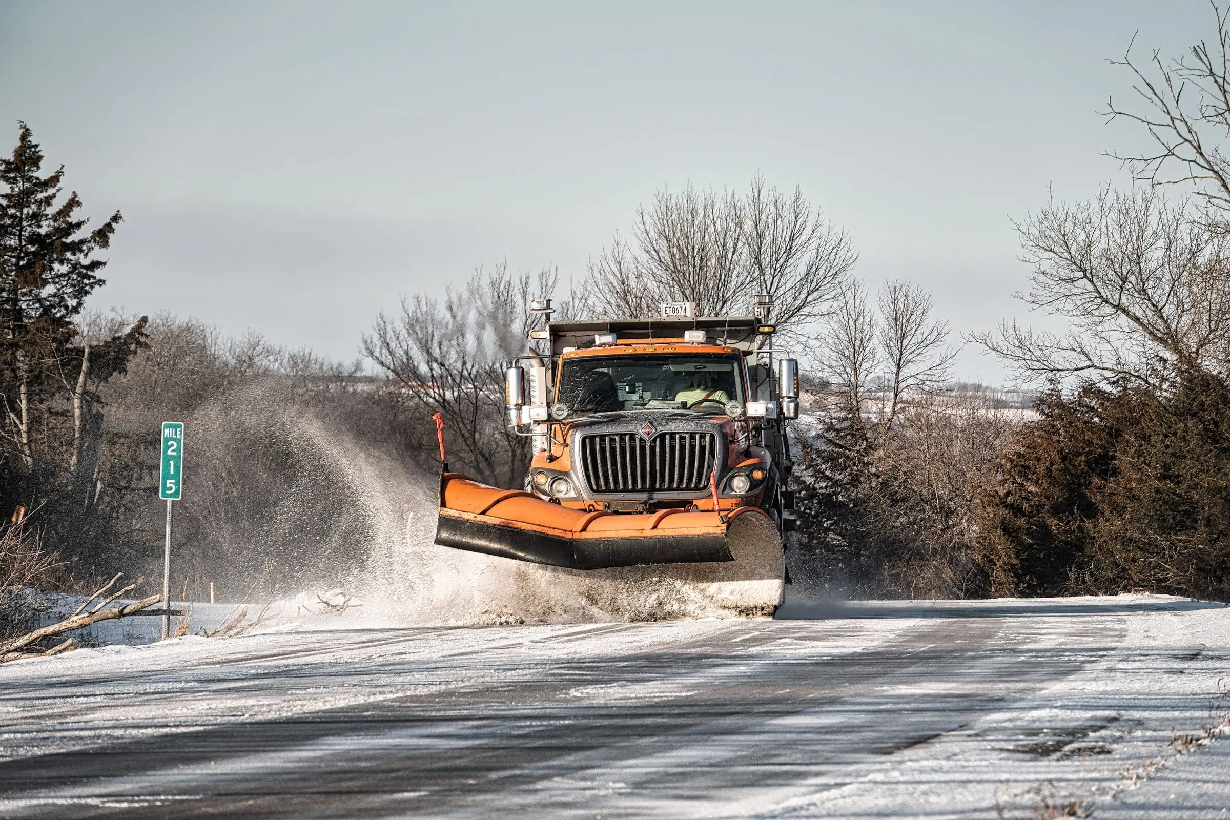 An orange highway plow truck with a front blade throwing snow off a rural two-lane road, bare trees and junipers either side and a green 'MILE 215' marker at left.
