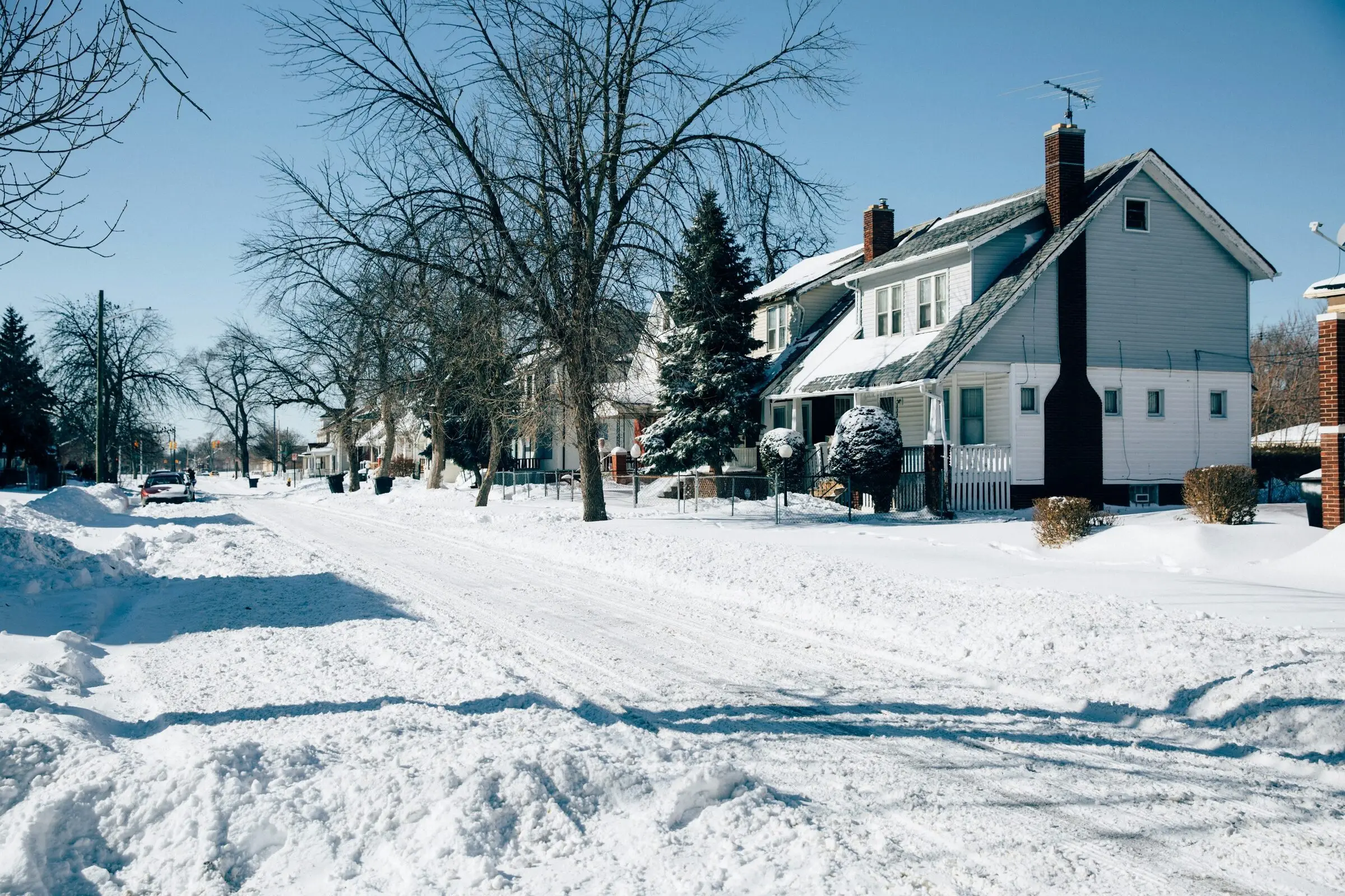A residential street after a heavy snowfall: rutted snow on the road, snowbanks along the curb, bare trees and a white two-storey house with a brick chimney under a clear blue sky.