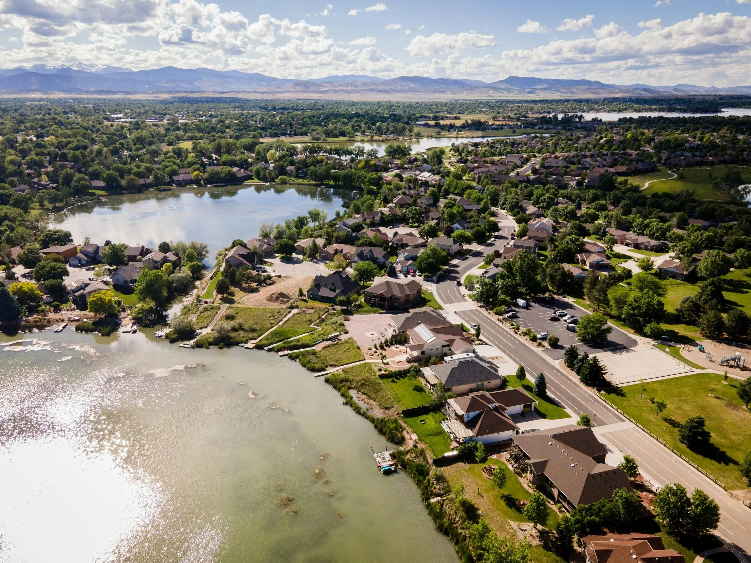 Aerial view of a lakeside residential neighbourhood with tree-lined streets, a park and playground, more lakes beyond and the Front Range foothills and snowy peaks on the horizon under scattered cumulus.