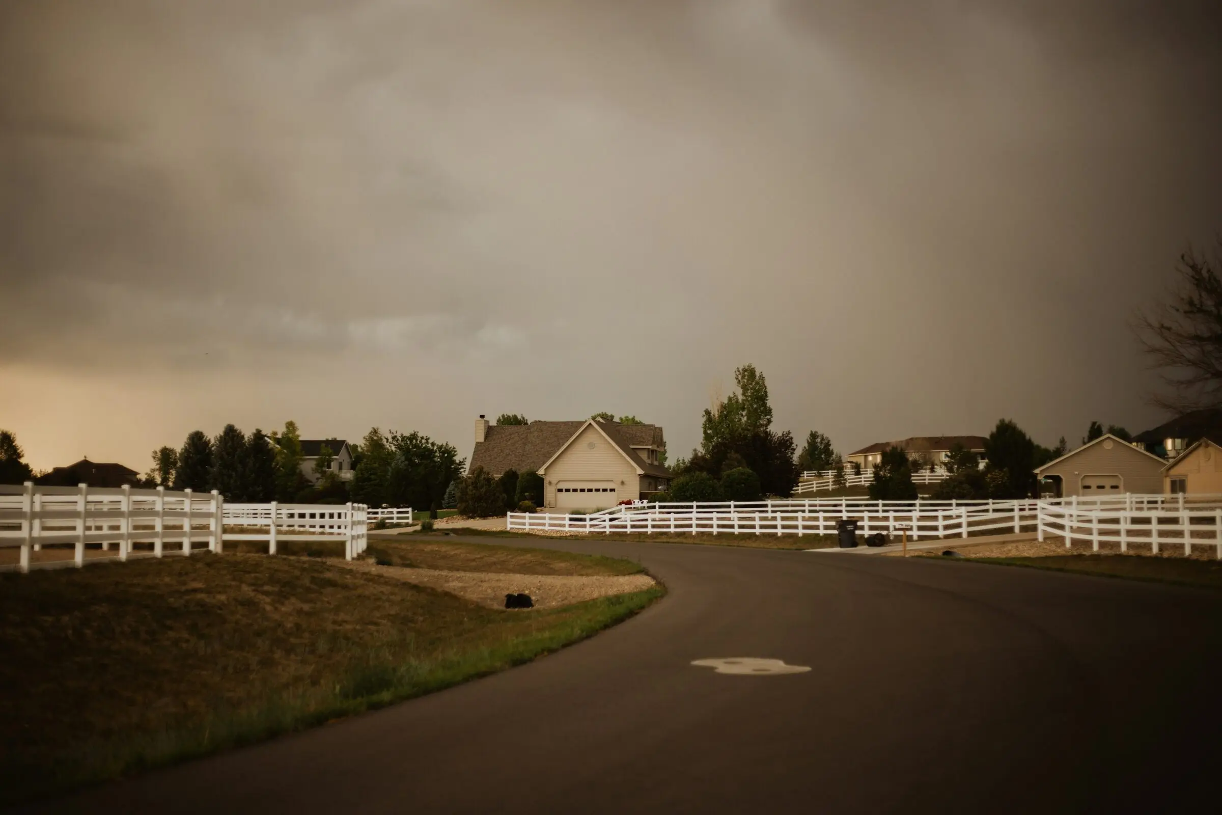 A curving residential lane past acreage homes with white rail fences, a tan house with an attached garage in the centre, under a dark brown-grey thunderstorm sky.