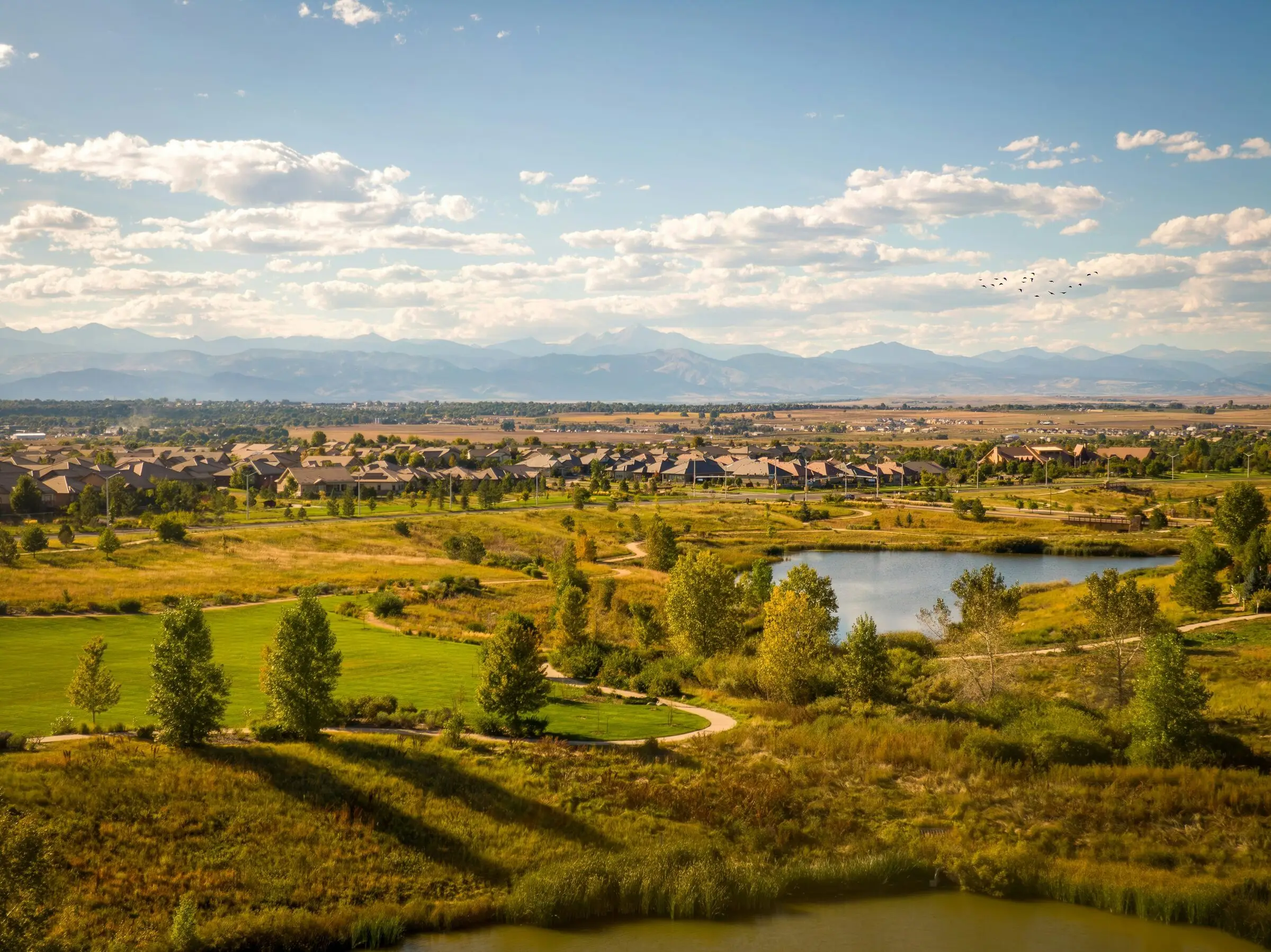 Elevated view over a park with a winding path, a pond, cottonwoods and green lawn, a subdivision of tan-roofed houses beyond and the Front Range with Longs Peak on the horizon under puffy clouds.