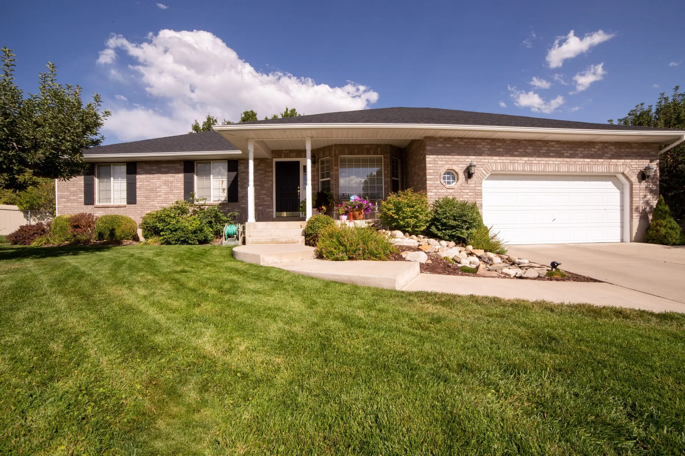 A tan brick ranch house with a white two-car garage, a concrete walk and a rock-and-shrub bed, fronted by a green lawn with mowing stripes under a deep blue sky with cumulus clouds.