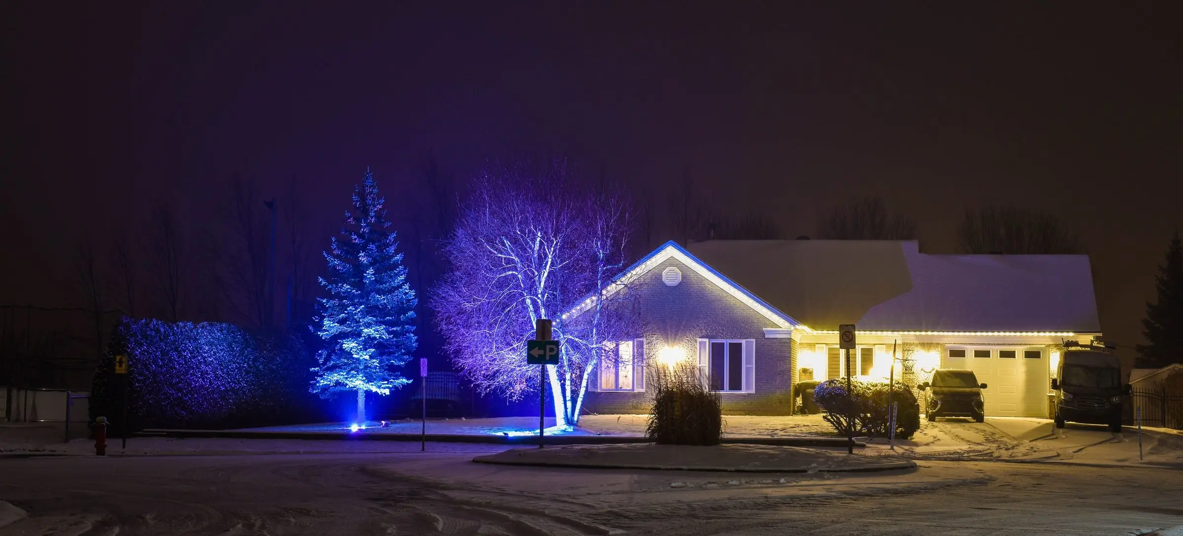 A single-storey house at night after light snowfall, warm white lights along the roof and eaves, a spruce and a birch lit in blue and violet uplight, a car and a van in the driveway, and a quiet snowy cul-de-sac in front.